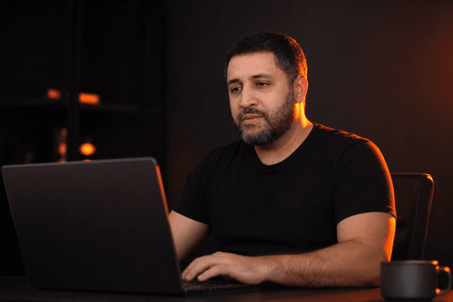 Danilo Gato sentado à mesa, trabalhando no notebook em um ambiente escuro com iluminação laranja