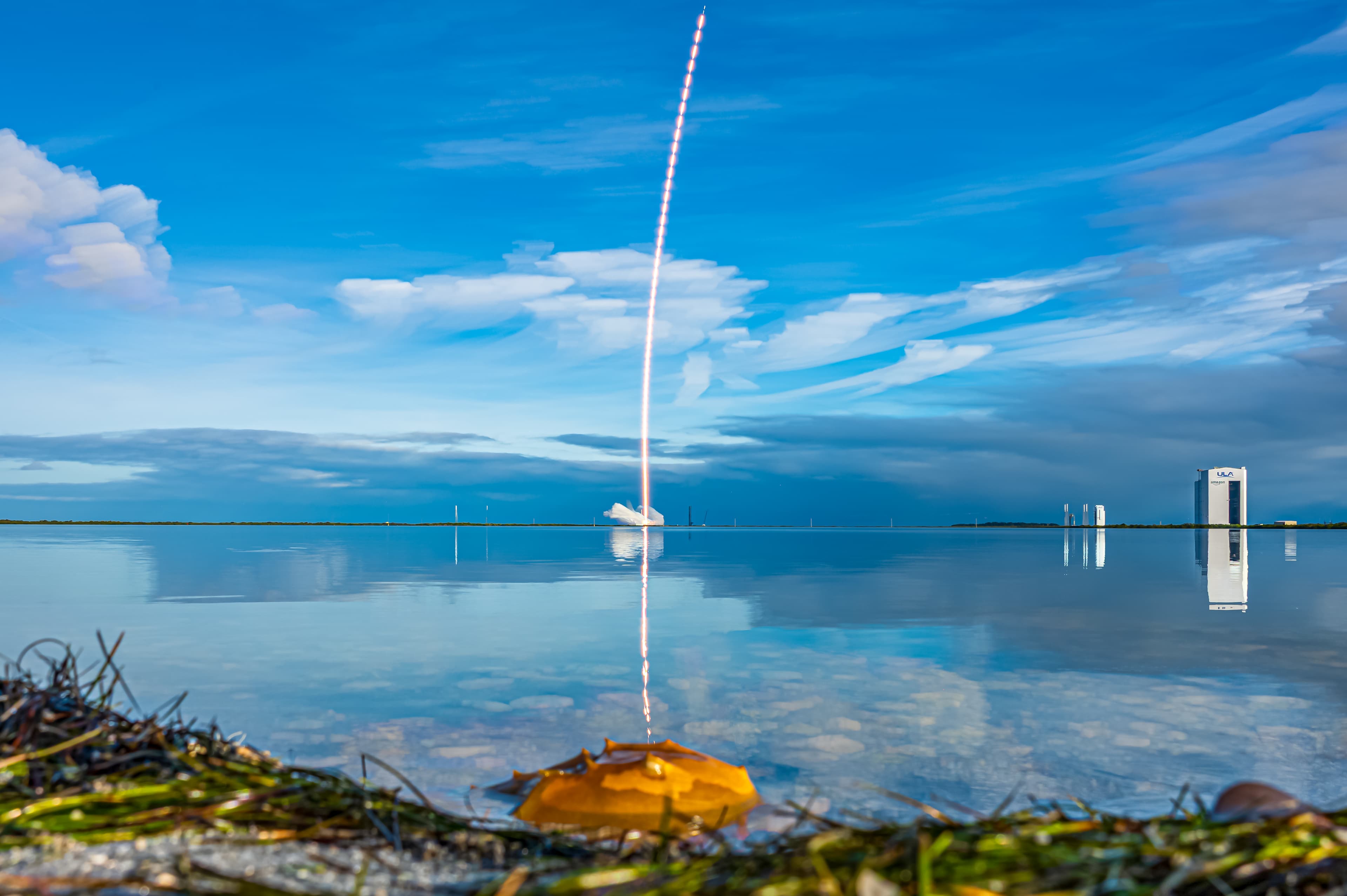 Trilha de luz de um Falcon 9 da SpaceX refletida em lago, céu azul com nuvens ao fundo