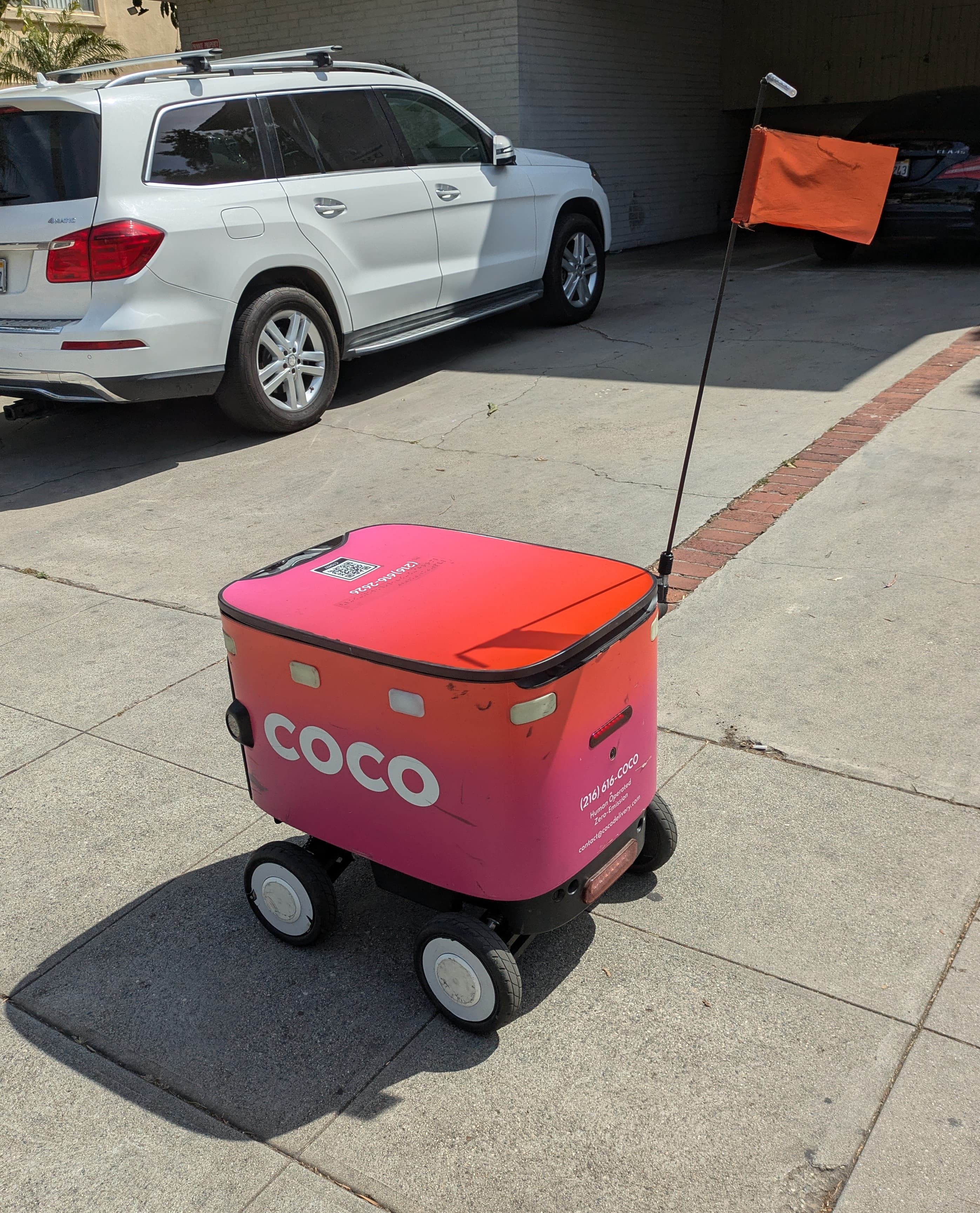 Robô de entrega Coco Robotics em calçada urbana, bandeira laranja visível