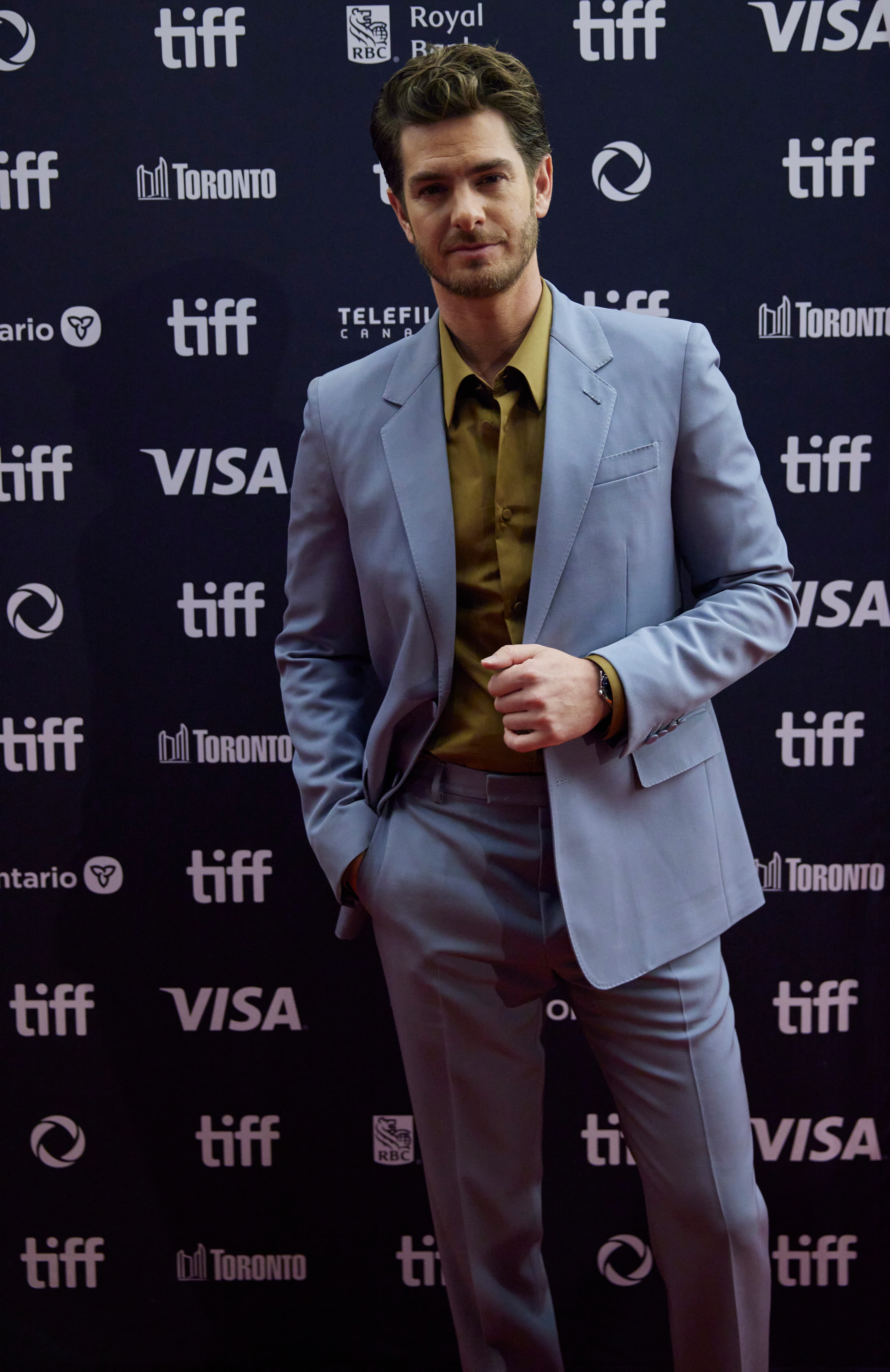 Andrew Garfield em evento de cinema, terno claro, em frente a painel de patrocinadores